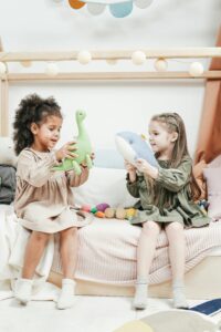 Children playing with stuffed animals. One girl holds a dinosaur while the other girl holds a shark.